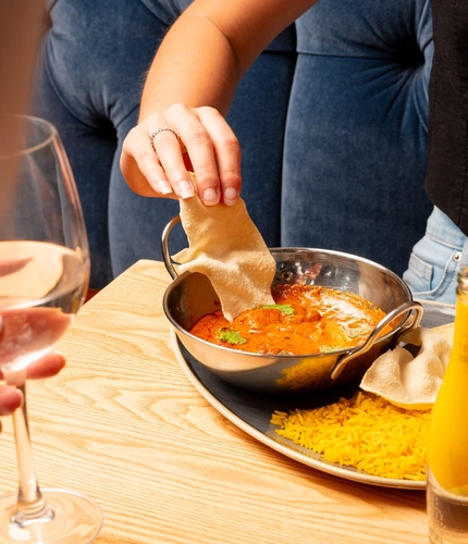 A hand dipping a poppadom into a balti dish containing curry on an indoor table.