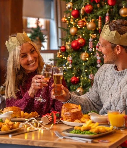 A festive Christmas meal is served at a dining table beside a decorated Christmas tree with red and gold baubles, candy cane ornaments and warm fairy lights. Four diners wear paper crowns from Christmas crackers while seated around the table. Two people raise glasses in a toast, while plates of traditional festive food, gravy boats, glasses of orange juice, Christmas crackers and string lights are arranged across the table. The setting is a warmly decorated restaurant with seasonal Christmas décor throughout.