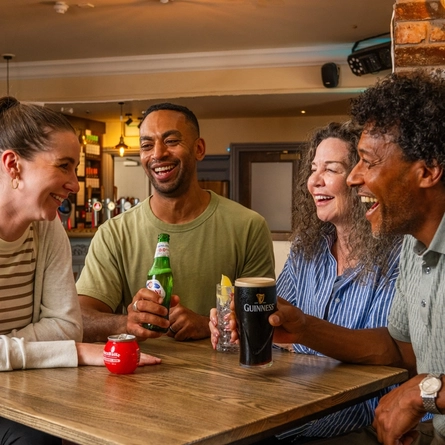 Four people gathered around a wooden table in a pub, socialising with a selection of drinks. The drinks include a pint of Guinness, a bottled alcohol-free Peroni, a mixed drink with ice and lemon, and a can of soft drink. Exposed brickwork, a stocked bar, warm pendant lighting and wooden furnishings create a welcoming pub atmosphere in the background.