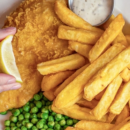A close up view of a plate of fish, chips, peas, and a small pot of sauce. A hand is squeezing a slice of lemon over the fish.