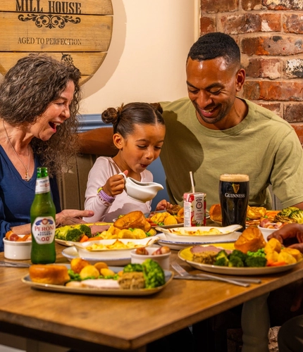 Three people seated around a wooden table in a pub, sharing Sunday roast meals. One person pours gravy from a small jug onto a plate of roast dinner with Yorkshire puddings, vegetables and potatoes. A bottle of alcohol-free beer, a pint of Guinness and a children's juice carton sit among the dishes. The background features blue panelled walls, exposed brickwork and a wooden barrel-style sign reading “The Mill House”.