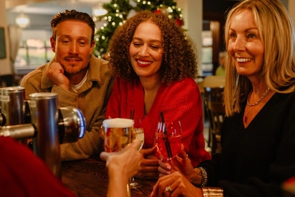 Three people sit together at a bar in a festively decorated pub, holding drinks and looking towards someone off camera. Glasses of rosé wine, a pint of beer and a mixed drink are arranged on the wooden bar, while a Christmas tree with warm fairy lights glows in the background, creating a cosy seasonal atmosphere.