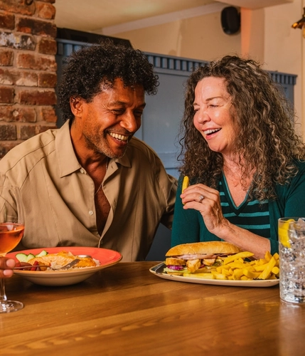 Two people seated at a wooden table in a pub, enjoying lunch and drinks. One plate holds a sandwich with chips, while another contains a salad with sliced vegetables. A glass of rosé wine and a clear drink with ice and lemon are on the table. Exposed brickwork, blue panelled dividers, leather seating and warm pendant lighting create a cosy pub interior, with a window overlooking greenery in the background.