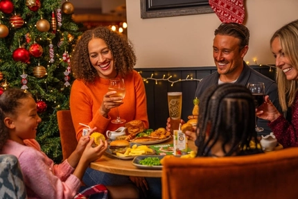 A group of diners gather around a festive table beside a decorated Christmas tree with red and gold baubles, candy cane ornaments and warm fairy lights. Plates of food, drinks and Christmas-themed table decorations are spread across the table. One diner holds a glass of white wine, another holds a pint of beer, and two children are seated nearby with food and drinks. The restaurant is decorated with seasonal lighting and Christmas décor, creating a warm festive setting.