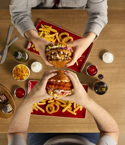 The hands of two people seated at a wooden table each holding a burger.  On teh table in front of each person are plates of fries and onion rings.