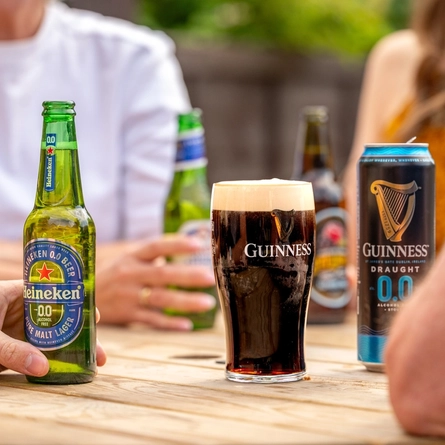 A wooden table with a can and pint of Guinness 0.0 Draught in the centre. Surrounding the table are the blurred out torsos of four people, each holding different drinks. 