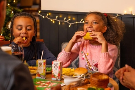 Two children sit at a festive dining table eating from plates of food, with one holding a piece of corn on the cob. The table is set with burgers, chips, vegetables, gravy, fruit juice cartons and drinks. Warm white fairy lights decorate the wall behind them, while a decorated Christmas tree with red and gold baubles and candy cane ornaments is partially visible to the side. Other diners are seated around the table in a cosy Christmas-themed restaurant setting.