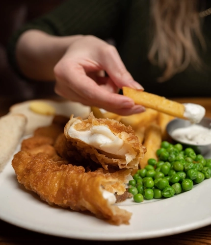 A close up view of a plate of fish, chips, peas, and a small pot of sauce, sitting on a wooden table. A hand is dipping one of the chips into the sauce.