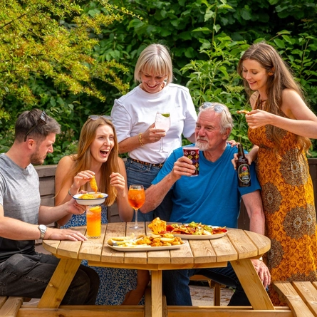 A mixed group of people seated and stood around a wooden table, in a beer garden, enjoying a variety of sharers and drinks.