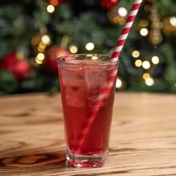 A close up view of a festive mocktail served in a highball glass with ice and a candy cane pattern straw. The glass sits on a wooden table and a Christmas tree is visible in the background.