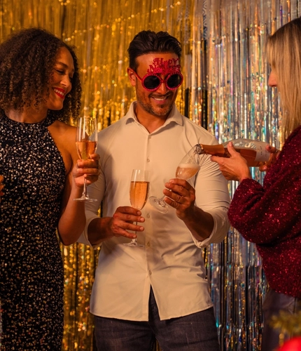 Four people gather in front of silver and gold metallic party backdrops during a festive New Year’s celebration. Three adults hold champagne flutes while one person wearing novelty red “Happy New Year” glasses stands in the centre. A child wearing a sequinned outfit stands nearby. A decorated Christmas tree with red and gold baubles and warm fairy lights is visible in the foreground, adding seasonal detail to the party setting.