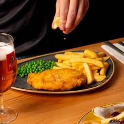 A hand squeezing a lemon wedge over a plated portion of fish and chips, at an indoor table.